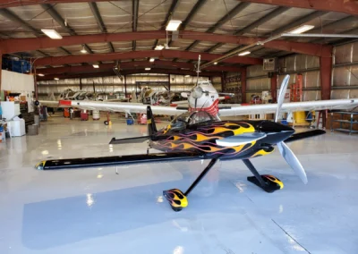 The interior of a bright hangar featuring a sleek black aerobatic plane with yellow and orange flame decals in the foreground, with several crop dusters parked behind it.